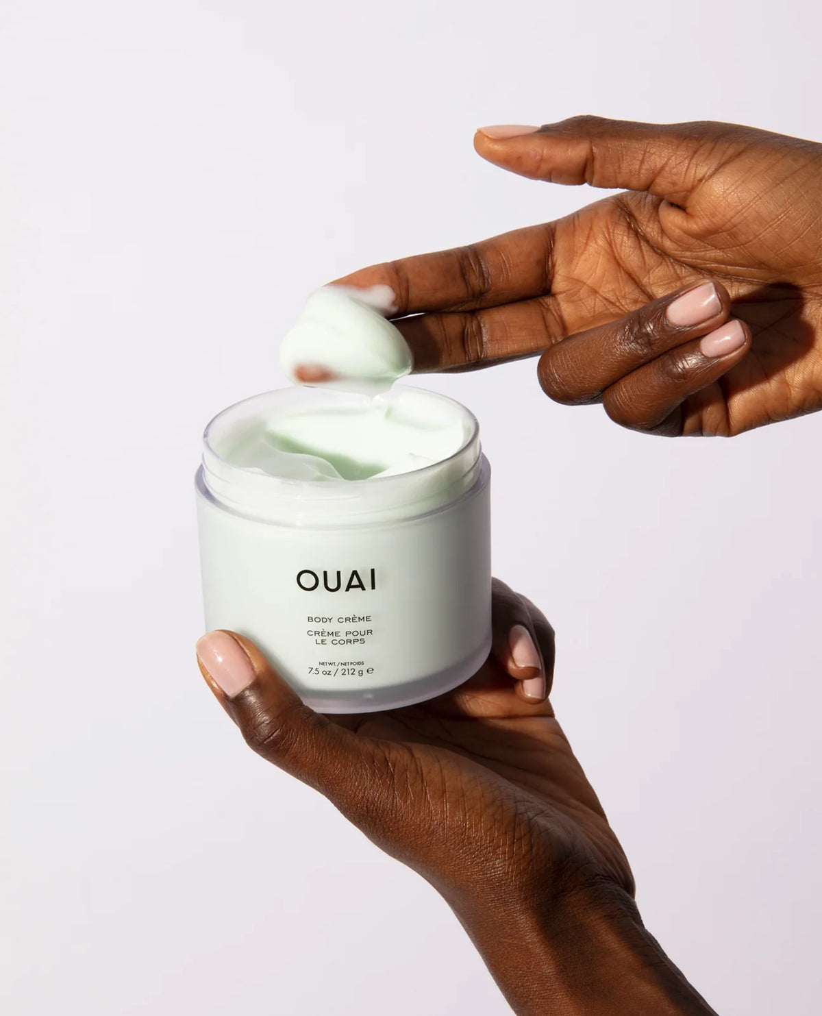 A person holds an open jar of OUAI body lotion while another hand scoops pale green cream from the jar against a clean white background.